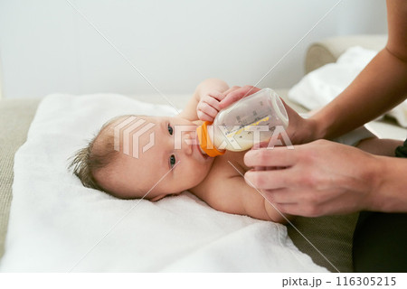 Hands of mother milk and feeding baby on sofa for growth development, nutrition or health. Drinking, woman parent and infant child with bottle for formula, wellness or bonding with love in home Hands of mother milk and feeding baby on sofa for growth development, nutrition or health. Drinking, woman parent and infant child with bottle for formula, wellness or bonding with love in home 116305215