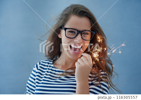 Happiness, portrait and woman in studio with sparklers isolated on blue background for celebration. Light, glasses and fun birthday event or new year with glow, vision and fireworks with confidence. Happiness, portrait and woman in studio with sparklers isolated on blue background for celebration. Light, glasses and fun birthday event or new year with glow, vision and fireworks with confidence. 116305557