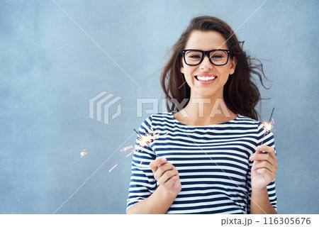 Portrait, studio and happy woman with sparklers for light with celebration isolated on blue background. Smile, glasses and new years party or fun birthday event with confidence, glow and fireworks. 116305676