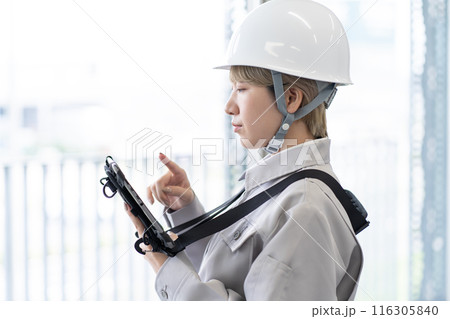 ヘルメットを被り作業服でタブレット端末を操作する金髪ショートヘアの若い女性作業員 116305840