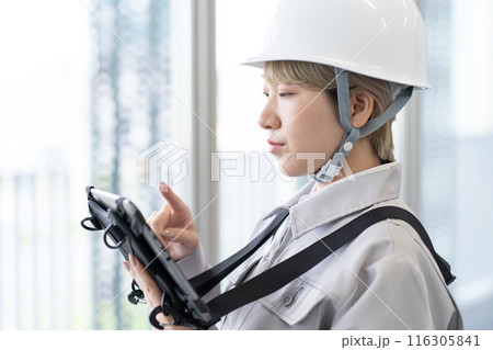 ヘルメットを被り作業服でタブレット端末を操作する金髪ショートヘアの若い女性作業員 116305841