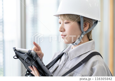 ヘルメットを被り作業服でタブレット端末を操作する金髪ショートヘアの若い女性作業員 116305842