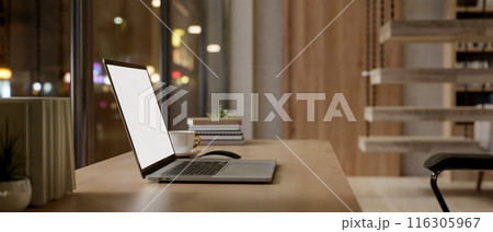 A close-up side view image of a laptop computer mockup on a wooden table in a modern home at night. 116305967