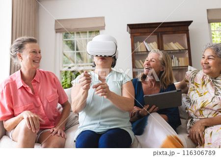 Diverse senior female friends trying on VR headset at home, laughing together 116306789