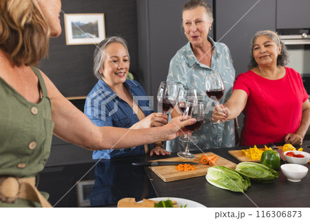 Diverse senior female friends enjoying wine at home in kitchen 116306873