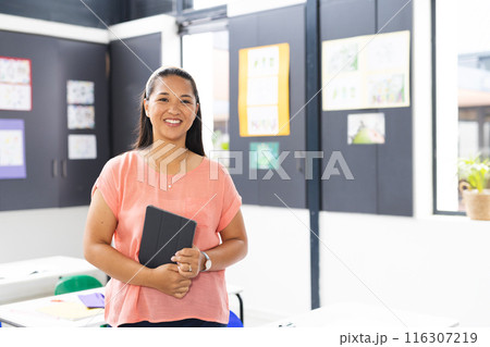 In school, young biracial female teacher holding a tablet, smiling, with copy space, in classroom 116307219