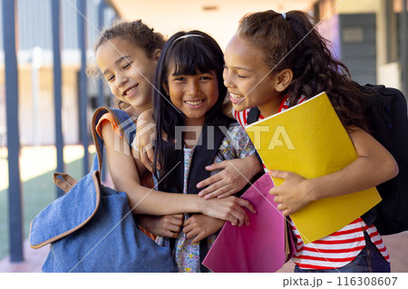 Three biracial girls are embracing outside a school, smiling with notebooks in hand 116308607