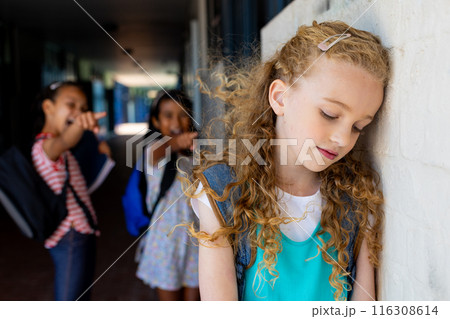 Caucasian girl leans on wall as two biracial girls whisper and engage in bull at school Caucasian girl leans on wall as two biracial girls whisper and engage in bull at school 116308614
