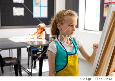 Caucasian girl with blonde hair paints on an easel, wearing a blue apron in school 116308638