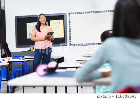 In school, young biracial female teacher holding tablet stands smiling in classroom In school, young biracial female teacher holding tablet stands smiling in classroom 116308645