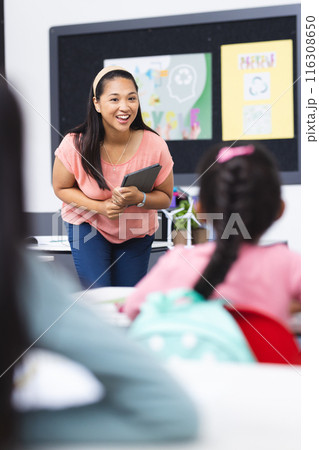 In school, young biracial female teacher holding a tablet stands before students 116308650
