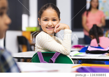 Young biracial girl with brown hair is sitting, smiling at her desk in classroom Young biracial girl with brown hair is sitting, smiling at her desk in classroom 116308691