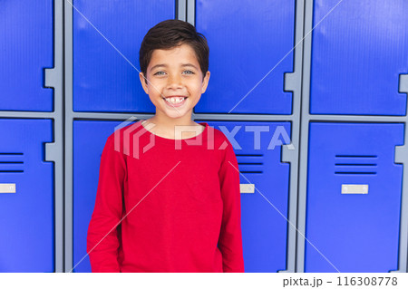 In school, young biracial male student standing in front of blue lockers outdoors, smiling 116308778