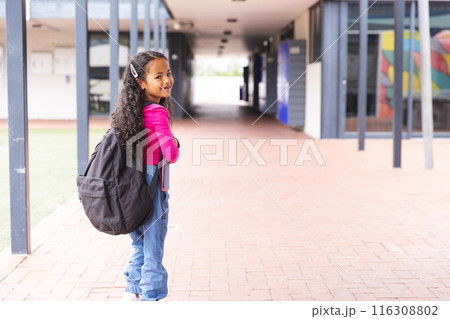 In school, a young biracial girl wearing a backpack standing in a hallway outdoors with copy space 116308802