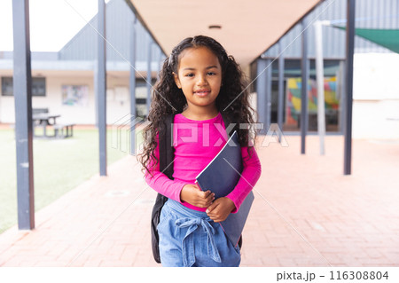 In school, young biracial girl holding a book, standing outside outdoors 116308804