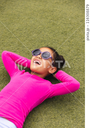 In school, outdoors, a biracial young girl wearing sunglasses is lying on grass, laughing In school, outdoors, a biracial young girl wearing sunglasses is lying on grass, laughing 116308879