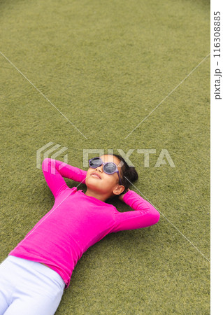 In school, outdoors, biracial young girl wearing sunglasses lying on grass with copy space 116308885