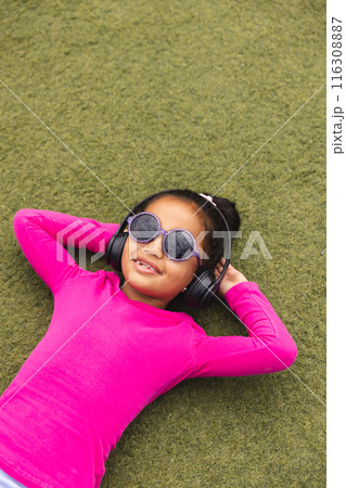 In school, young biracial girl is lying on grass outdoors, wearing headphones 116308887