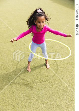 In school, outdoors, a biracial young girl is playing with a yellow hula hoop 116308904