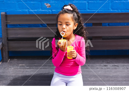 In school, young biracial girl wearing pink blowing bubbles outdoors 116308907