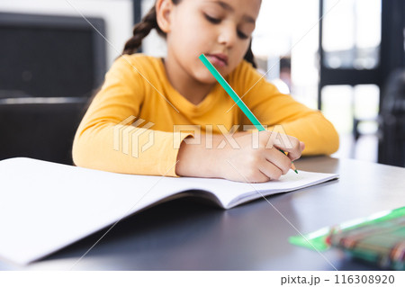 In school, young biracial girl wearing a yellow top writing in a notebook in the classroom 116308920