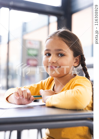 In school, young biracial girl holding a pencil, sitting at a desk in a classroom 116308929