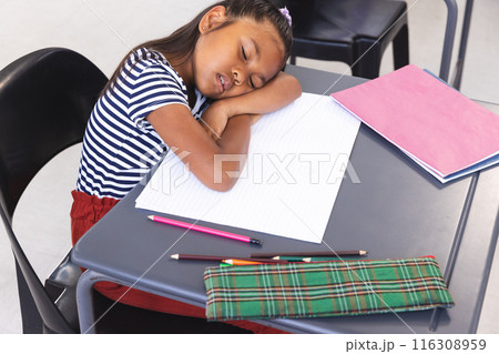 In school, young biracial girl wearing a striped shirt resting head on desk in classroom 116308959