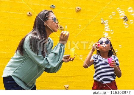 A woman and young biracial girl are blowing bubbles outdoors against a yellow wall 116308977
