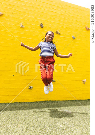 In school, outdoors, a biracial young girl is jumping high near a climbing wall 116308982