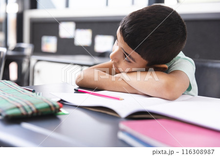 In school, young biracial boy resting head on arms in classroom, looking at notebook 116308987