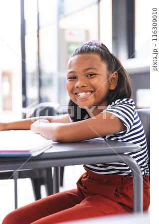 In school, young biracial girl with a bright smile sitting at a desk in a classroom 116309010