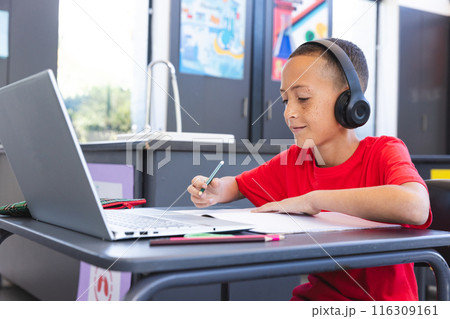 Biracial boy studies intently in a school classroom using a laptop, with copy space 116309161