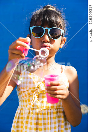 Biracial girl wearing sunglasses and a sundress blows bubbles outdoors 116309317