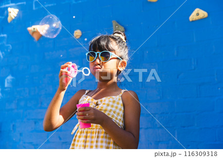 Biracial girl wearing sunglasses and a sundress enjoys blowing bubbles outdoors, with copy space 116309318