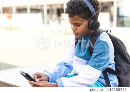 Biracial boy enjoys music on his phone outdoors at school, with copy space 116309433