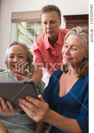 Diverse senior female friends sharing tablet at home, all smiling together Diverse senior female friends sharing tablet at home, all smiling together 116309440