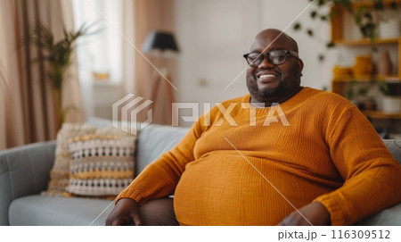 Fat lazy african american man on sofa at home wearing orange warm sweater. Disappointment and gain. 116309512