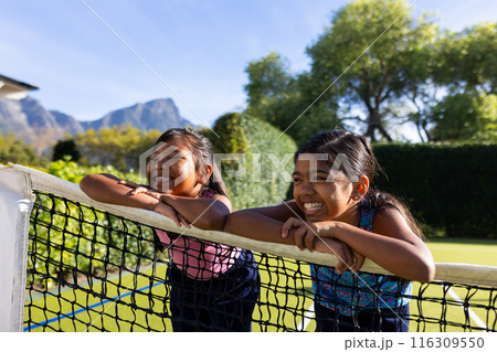 Outdoors, two biracial young sisters leaning on tennis net, smiling and having fun 116309550