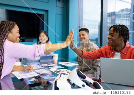 Young diverse Business colleagues high-five and smile around a conference table in a startup Young diverse Business colleagues high-five and smile around a conference table in a startup 116310158