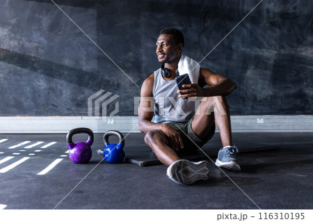 Fit African American man sitting on gym floor with smartphone after exercising, copy space Fit African American man sitting on gym floor with smartphone after exercising, copy space 116310195