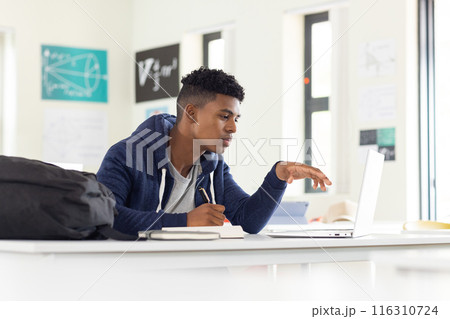 In school, African American male student using laptop and writing in notebook, studying 116310724