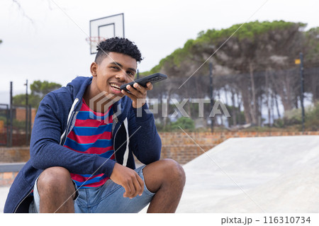 African American teenage boy using smartphone voice command, sitting at skatepark, copy space 116310734