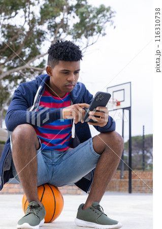Sitting on basketball, teenage boy using smartphone at outdoor court, focusing intently 116310738