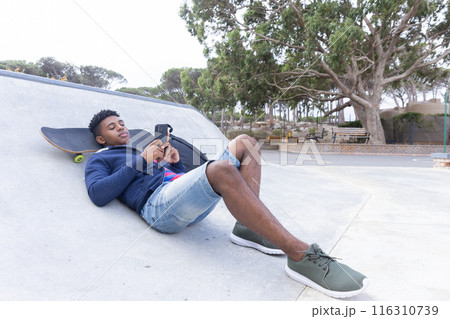 Relaxing at skatepark, African American teenage boy using smartphone, skateboard beside 116310739