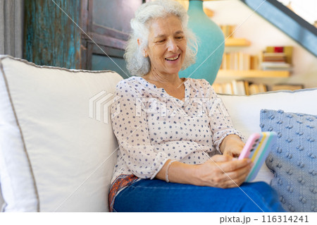 Senior woman smiling and using smartphone while relaxing on couch at home 116314241
