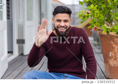 Smiling man waving hand while sitting on wooden deck at home, on video call 116314311