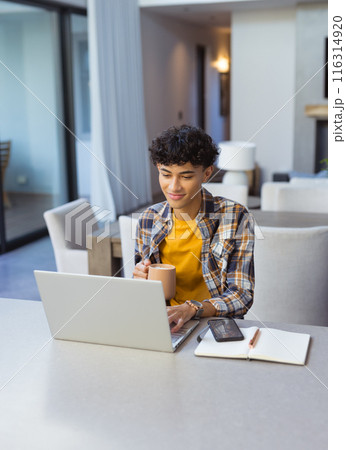 Working from home, teenage boy using laptop and holding coffee mug at table 116314920