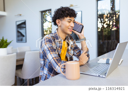 Talking on smartphone, teenage boy using laptop and smiling at home 116314921
