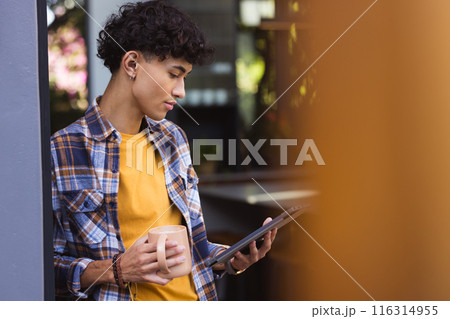 Holding mug, teenage boy reading on tablet, standing in living room, copy space 116314955
