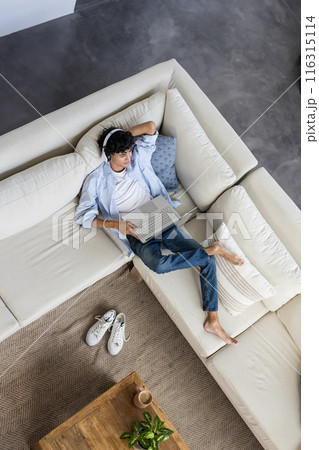 Relaxing on sofa, young man using laptop and wearing headphones at home 116315114